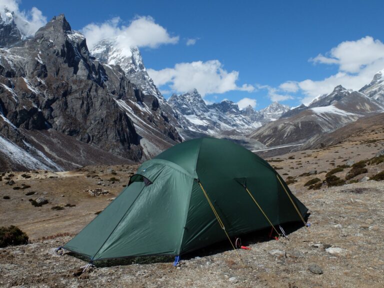 A single green expedition tent pitched on a rocky alpine plain with sparse shrubs, set against towering jagged snow‑tipped mountains under a bright blue sky with scattered clouds.