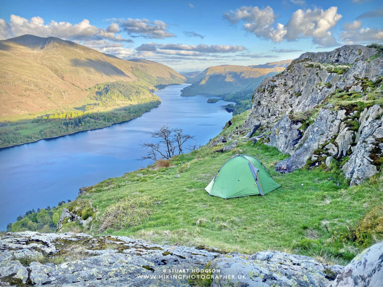 Green backpacking tent pitched on a grassy cliff ledge beside rocky outcrops, overlooking a long blue lake winding between sunlit hills and a lone bare tree under a partly cloudy sky.