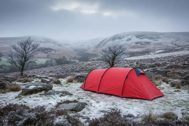 Red camping tent set against a snowy landscape, surrounded by rocky terrain and sparse vegetation, with misty mountains in the background. Ideal for winter outdoor adventures and camping enthusiasts.