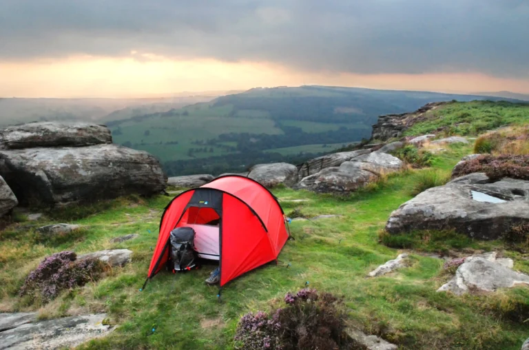Red camping tent set up on a grassy hillside with rocky outcrops, overlooking a scenic valley at sunset, ideal for outdoor adventure and nature exploration.