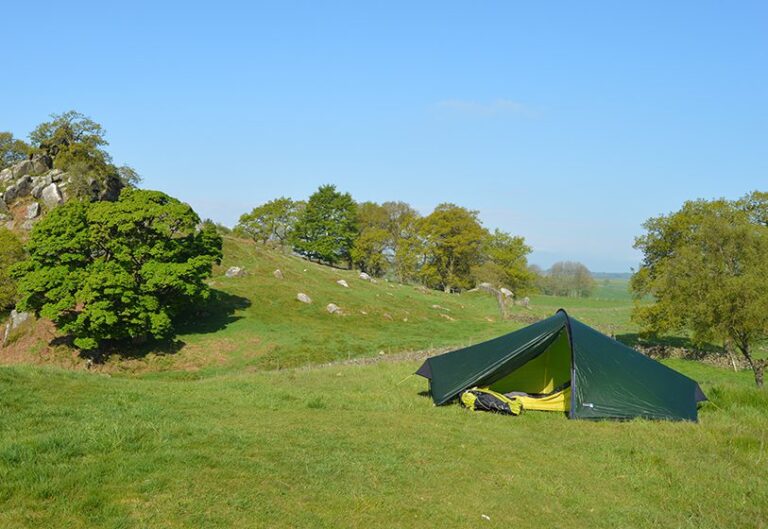 Green two-person tent with an open flap showing a yellow sleeping bag and backpack, pitched on a grassy hillside surrounded by scattered rocks, trees and a clear blue sky.