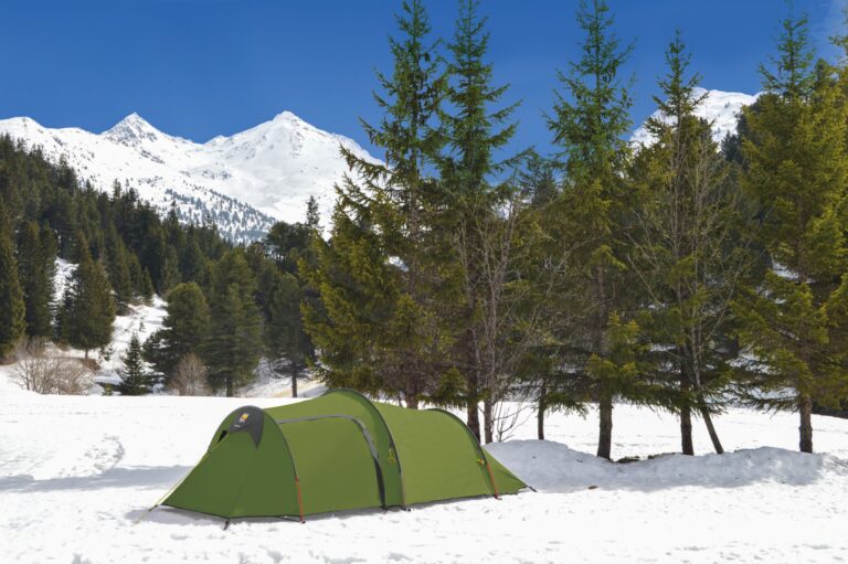 Long green tunnel tent pitched on a snowy clearing beside evergreen trees, with a dark conifer forest, snow-capped mountains and a clear blue sky in the background.