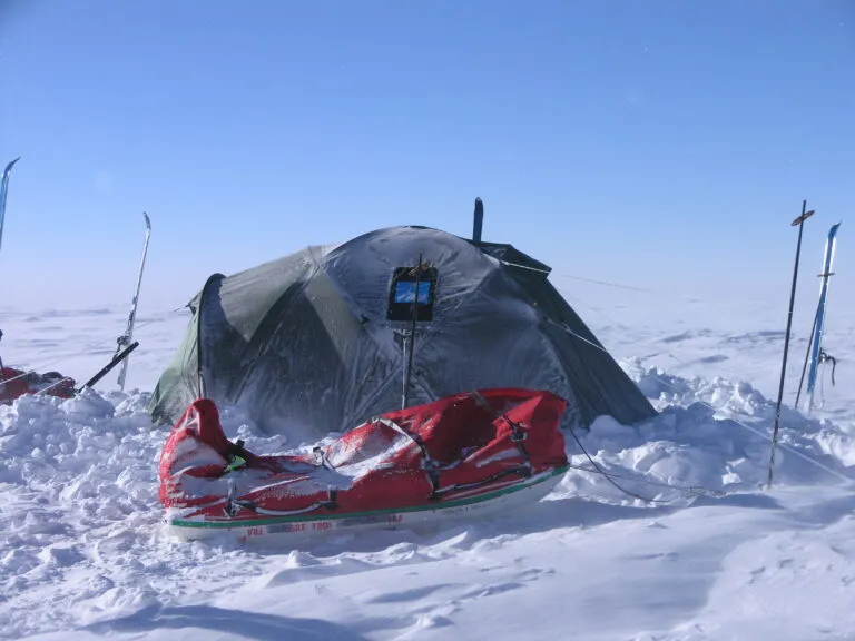 Tent set up in a snowy landscape, with a red sled in the foreground, showcasing a remote winter camping scene in extreme conditions.
