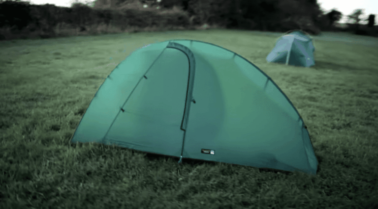 Green dome tent with a front zip door pitched on a grassy field, with a smaller tent and a tree line visible in the background.