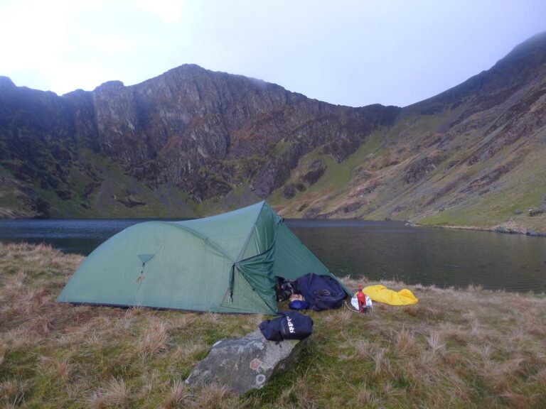 A green two-person tent pitched on grassy ground beside a calm mountain tarn, with backpacks and camping gear including a yellow dry bag and red fuel bottle beside it and steep rocky slopes rising around the lake.