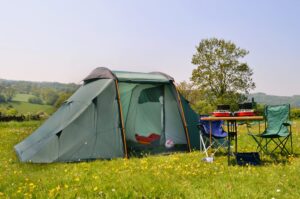 Green family tent pitched in a sunny wildflower meadow with a folding table and two camping chairs beside it, a double-burner stove and camping gear on the table, and rolling hills with a lone tree in the background.