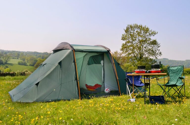 Green family tent pitched in a sunny wildflower meadow with a folding table and two camping chairs beside it, a double-burner stove and camping gear on the table, and rolling hills with a lone tree in the background.