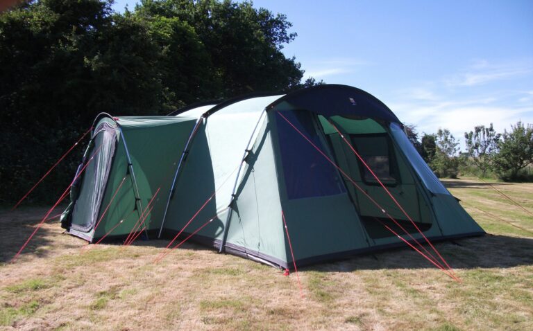 Large green multi-room tunnel tent with arched black poles and bright red guy lines staked into sun-browned grass, a partially open front doorway and trees under a clear blue sky.