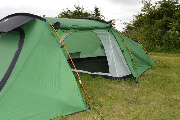 Green tunnel-style camping tent with orange support poles and an open front entrance revealing the inner sleeping area, pitched on grass with bushes and an overcast sky in the background.