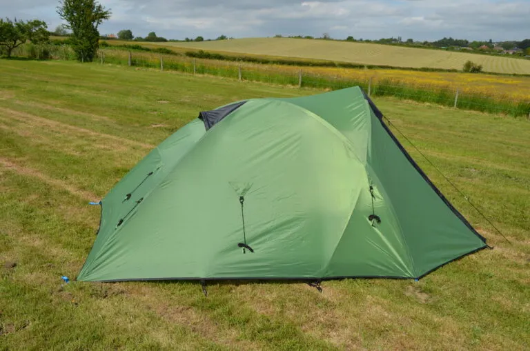Green camping tent set up on a grassy field with rolling hills and a cloudy sky in the background, ideal for outdoor adventures and camping trips.