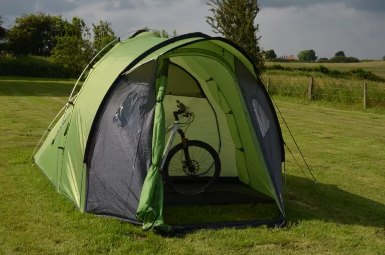 Green camping tent set up in a grassy area, featuring a bicycle inside, surrounded by trees and an open landscape. Ideal for outdoor enthusiasts and cyclists.