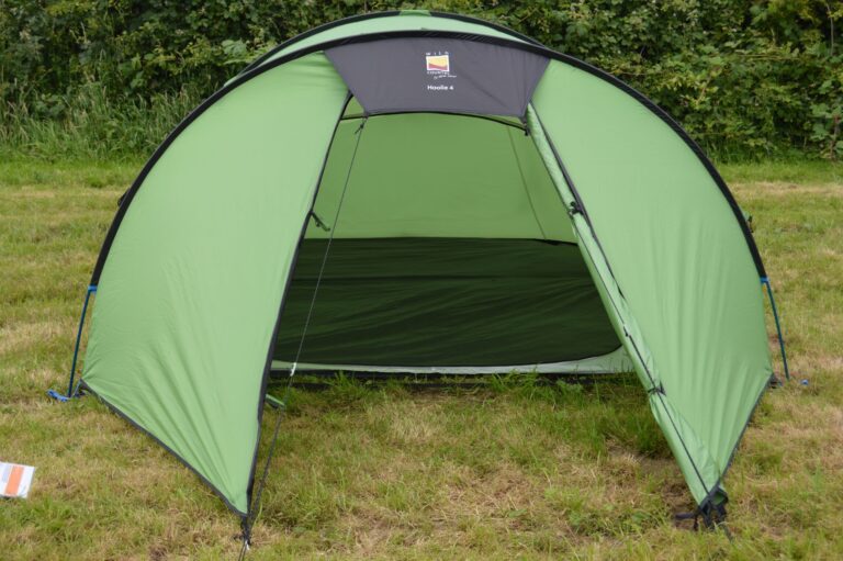 Green dome tent with its front open to reveal a dark sleeping area, pitched on a grassy field in front of a leafy hedge.