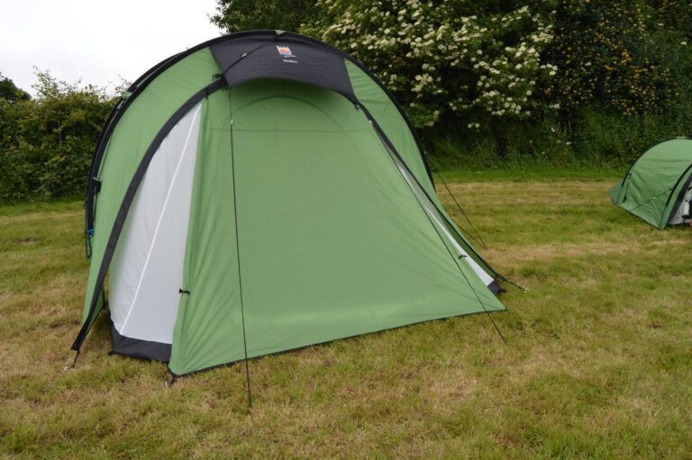 Large light-green dome camping tent with a small black canopy and black trim staked on a grassy field, with a hedgerow of white-flowering bushes behind and a smaller green tent partly visible at the right edge.