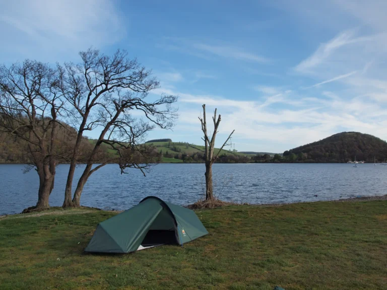 Tent set up by a serene lake surrounded by rolling hills and trees under a clear blue sky, ideal for camping and outdoor activities.