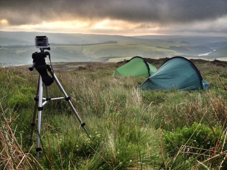 Tripod-mounted action camera in the foreground with two green tents pitched on grassy moorland overlooking rolling hills and a distant river beneath a cloudy dawn sky.