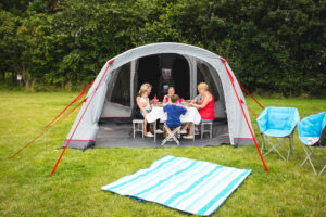 Adults and children sit around a small table inside a grey dome tent with red guy lines, sharing a meal while a striped blue-and-white picnic blanket lies on the grass in front and two blue folding camping chairs sit to the right against a hedge backdrop.