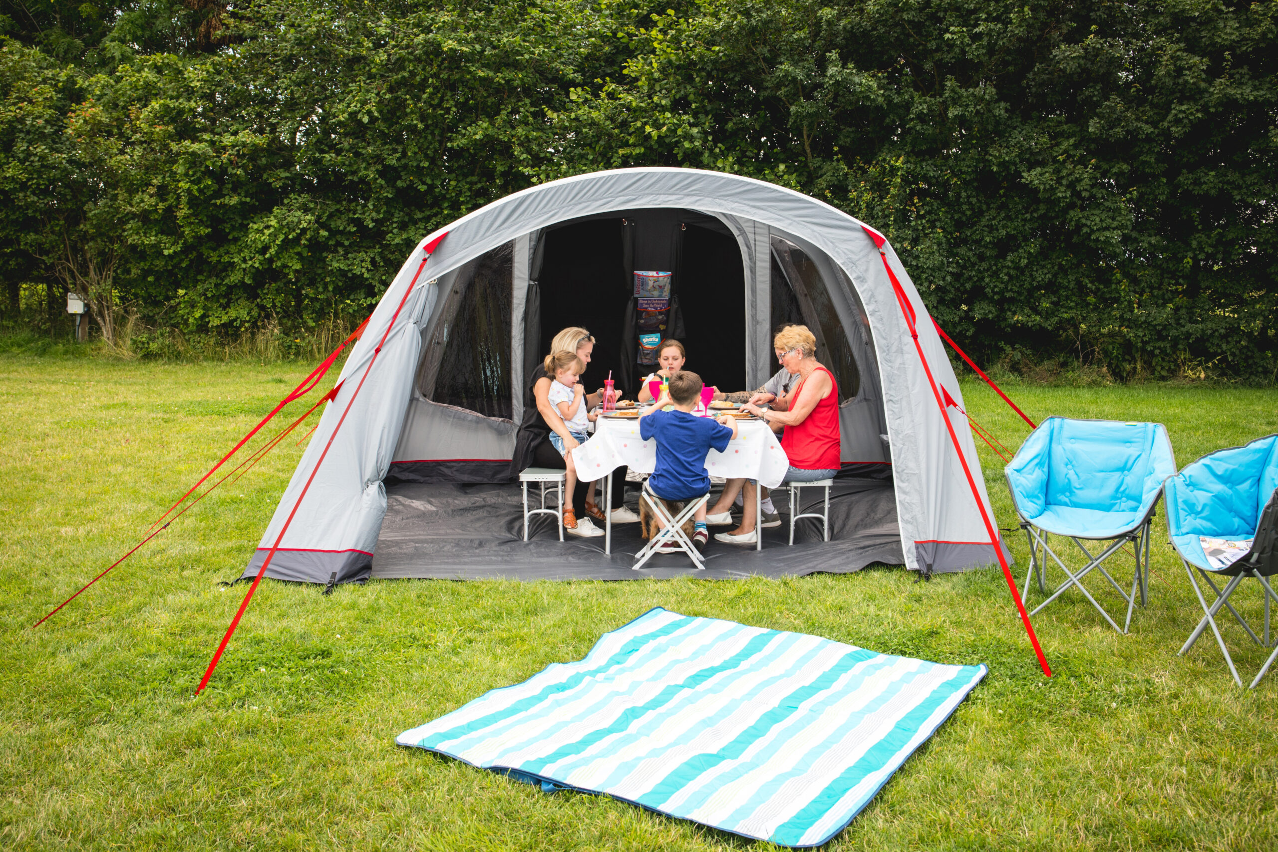 Adults and children sit around a small table inside a grey dome tent with red guy lines, sharing a meal while a striped blue-and-white picnic blanket lies on the grass in front and two blue folding camping chairs sit to the right against a hedge backdrop.