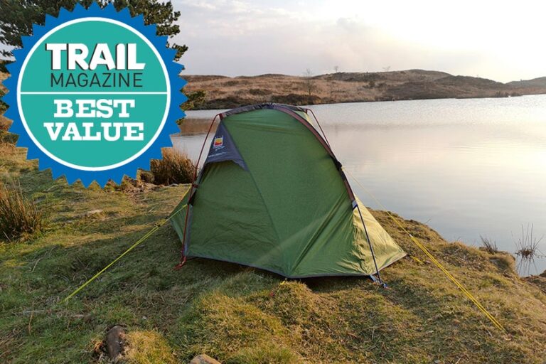 Green backpacking tent staked on a grassy lakeshore with yellow guy lines, calm water and rolling brown hills under a pale sky, and a circular 'Trail Magazine Best Value' badge overlaid in the top-left.