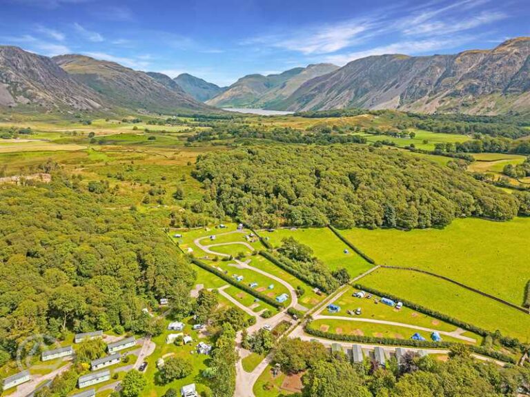 Aerial view of a lush green valley surrounded by mountains, featuring winding paths and patches of forest, with a clear blue sky overhead, showcasing the natural beauty of the landscape.