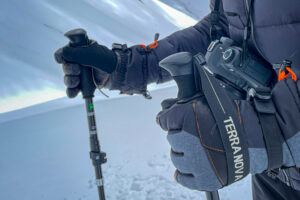 Close-up of gloved hands gripping trekking poles in snowy terrain, a DSLR camera hangs from a strap labelled TERRA NOVA and the person wears a dark insulated jacket.