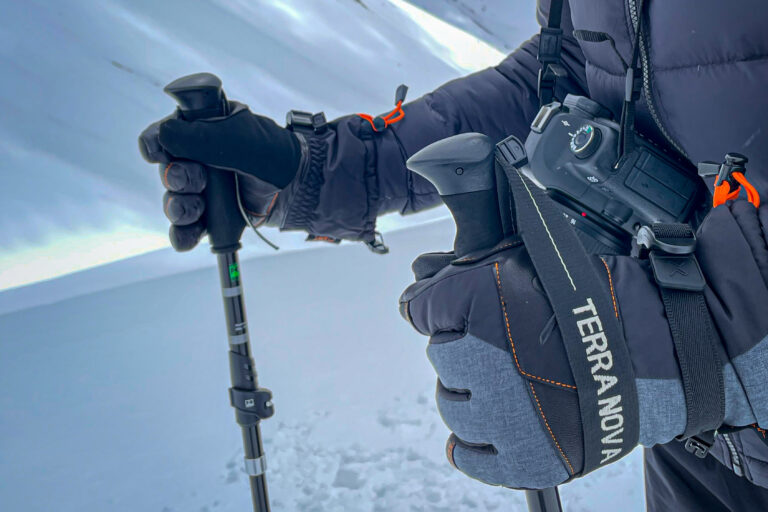 Close-up of gloved hands gripping trekking poles in snowy terrain, a DSLR camera hangs from a strap labelled TERRA NOVA and the person wears a dark insulated jacket.