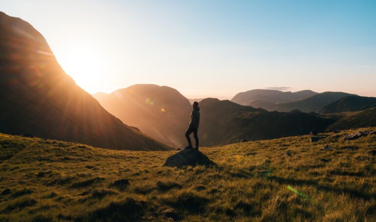 Silhouette of a person standing on a rock, overlooking a breathtaking mountain landscape at sunset, with warm sunlight illuminating the scene and highlighting the rolling hills and valleys.