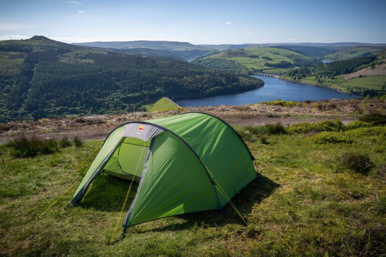 Bright green dome tent staked on a grassy hilltop with yellow guy lines, overlooking a blue reservoir and dam set among wooded hills and patchwork fields beneath a clear blue sky.