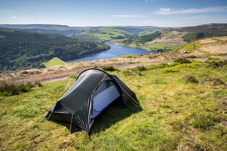 A small dark camping tent pitched on a grassy hillside overlooking a winding reservoir, forested valley and patchwork green hills beneath a clear blue sky.