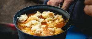Close-up of a dark metal mug of hot tomato-based soup topped with torn pieces of crusty bread, a spoon inside and a hand holding the handle, suggesting a rustic outdoor meal.