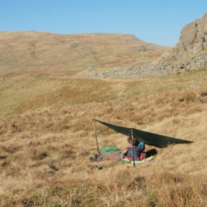 A person sitting under a makeshift shelter in a grassy, hilly landscape on a clear day, surrounded by rolling hills and rocky outcrops.