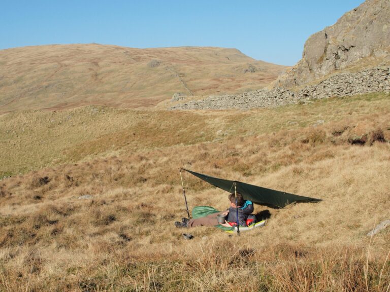A person sitting under a makeshift shelter in a grassy, hilly landscape on a clear day, surrounded by rolling hills and rocky outcrops.