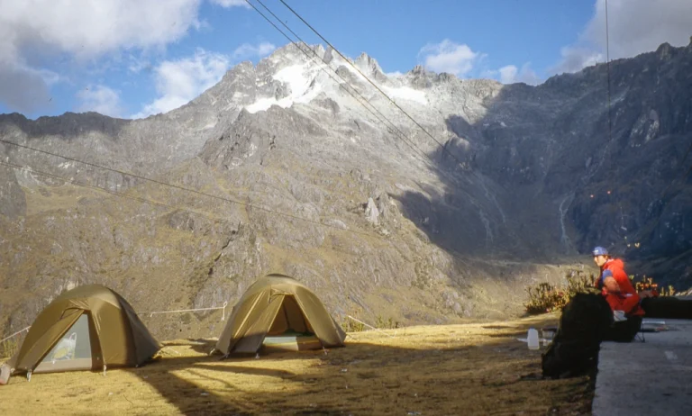 Two khaki tents pitched on a grassy alpine plateau beneath steep, snow-dusted jagged peaks with cable lines overhead, and a person in a red jacket sitting on the right.