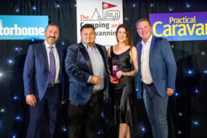 Group of four individuals at an award ceremony, celebrating achievements in the camping and caravanning industry. The backdrop features event branding and colorful signage.