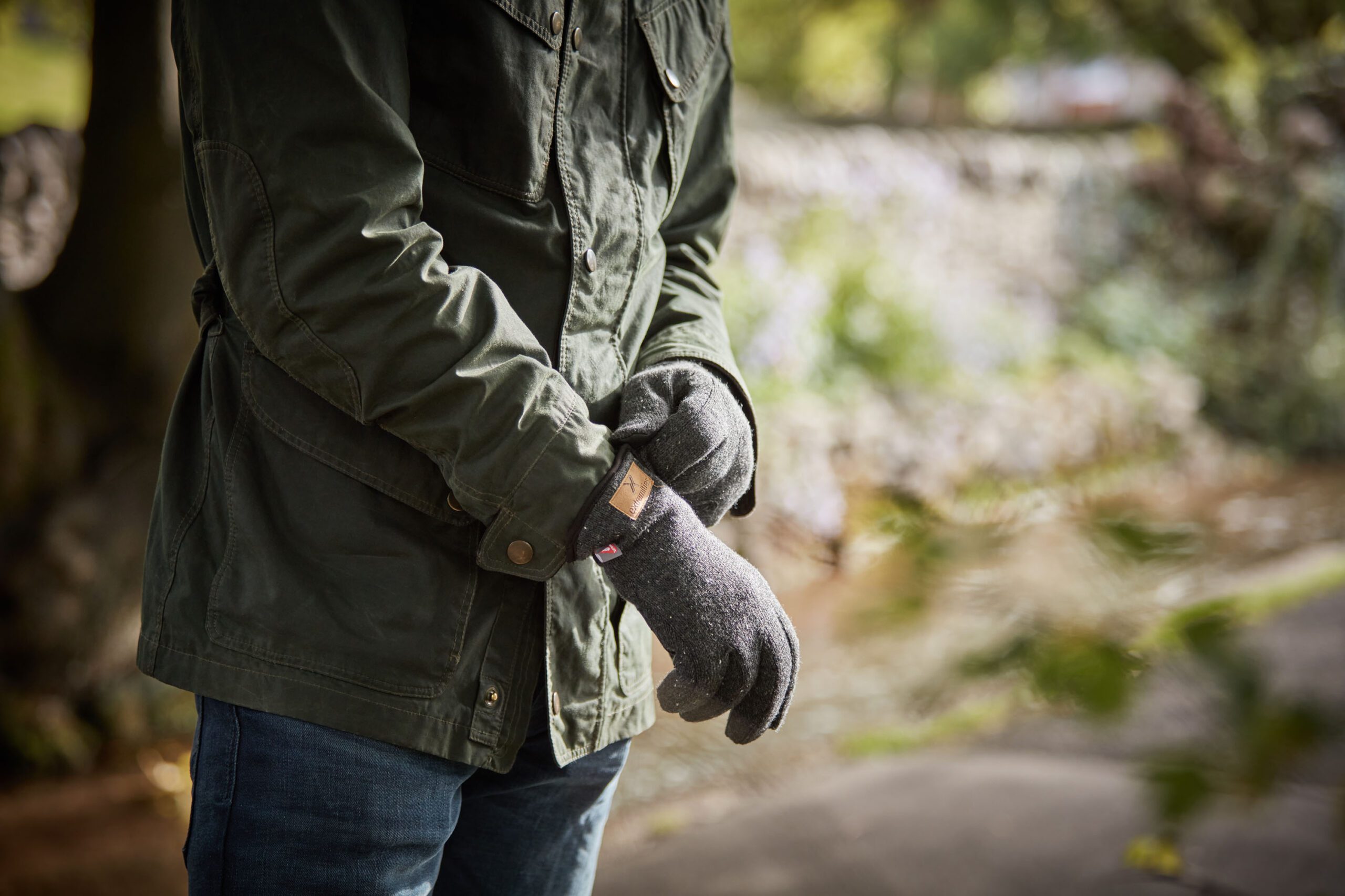 Person wearing a green jacket and black gloves, adjusting their wristwatch outdoors in a natural setting.