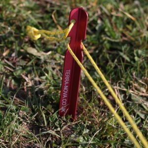 Red tent stake anchored in grass with yellow guy line attached, used for securing tents and outdoor gear.