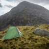 A small green tent with yellow guy lines pitched on grassy highland beside large grey boulders, with a steep rocky mountain filling the background under a cloudy sky.