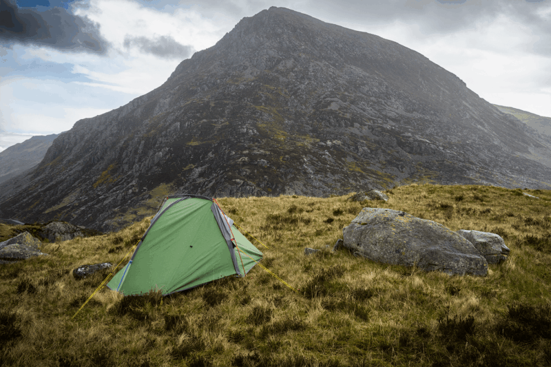 A small green tent with yellow guy lines pitched on grassy highland beside large grey boulders, with a steep rocky mountain filling the background under a cloudy sky.