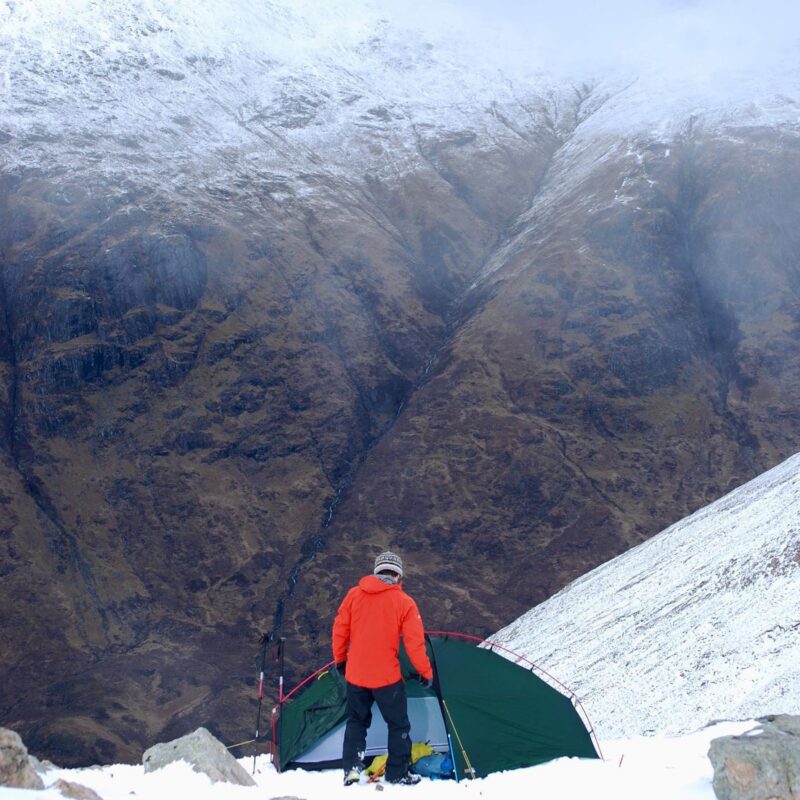 Person in an orange jacket and knit hat stands beside a green tent on a snowy mountainside, looking down into a steep, snow-dusted valley with rugged brown rock slopes.