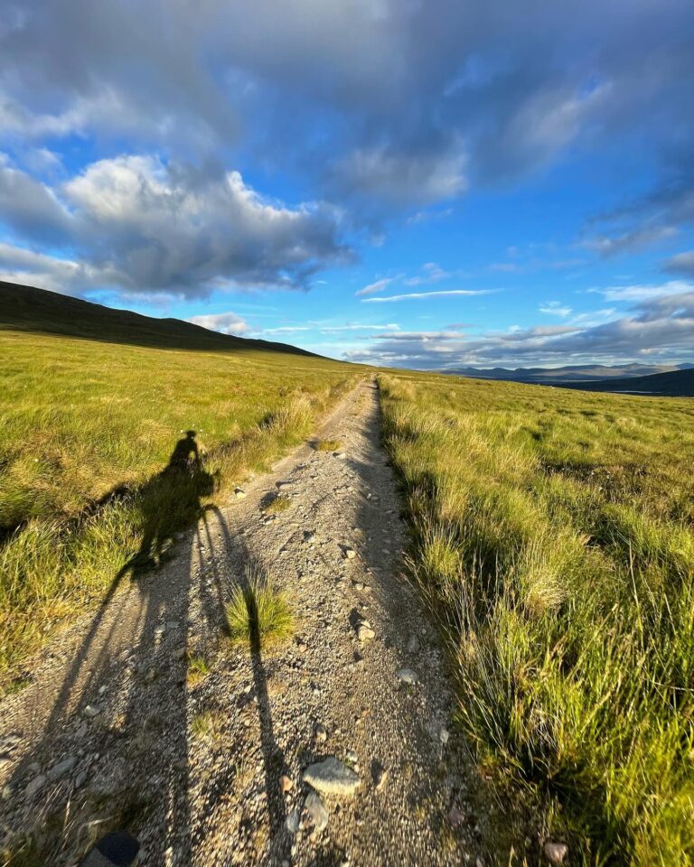 Bicyclist casting a shadow on a gravel path through a grassy landscape under a blue sky with clouds, ideal for outdoor cycling and adventure activities.