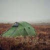 Green dome tent pitched in windswept brown moorland with yellow guy lines, surrounded by tall dry grass under a foggy, overcast sky.