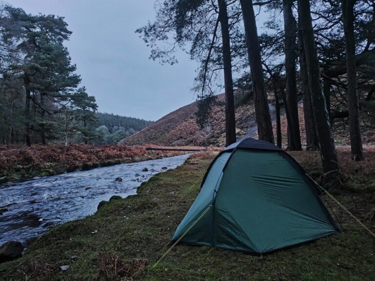 Tent set up by a river in a forested area during twilight, showcasing a peaceful camping scene surrounded by trees and hills.