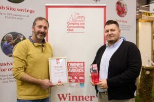 Two men smile in front of a Camping and Caravanning Club banner at an indoor event; the man on the left in a yellow pullover holds a framed 'Winner Terra Nova' certificate while the man on the right in a dark cardigan holds a red award trophy.