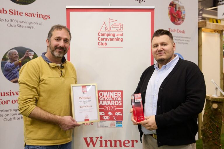Two men smile in front of a Camping and Caravanning Club banner at an indoor event; the man on the left in a yellow pullover holds a framed 'Winner Terra Nova' certificate while the man on the right in a dark cardigan holds a red award trophy.