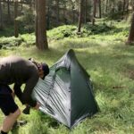 Person in a dark jacket and baseball cap bending to peg down a small green solo tent on long grass in a sunlit pine woodland with ferns and tall trees.