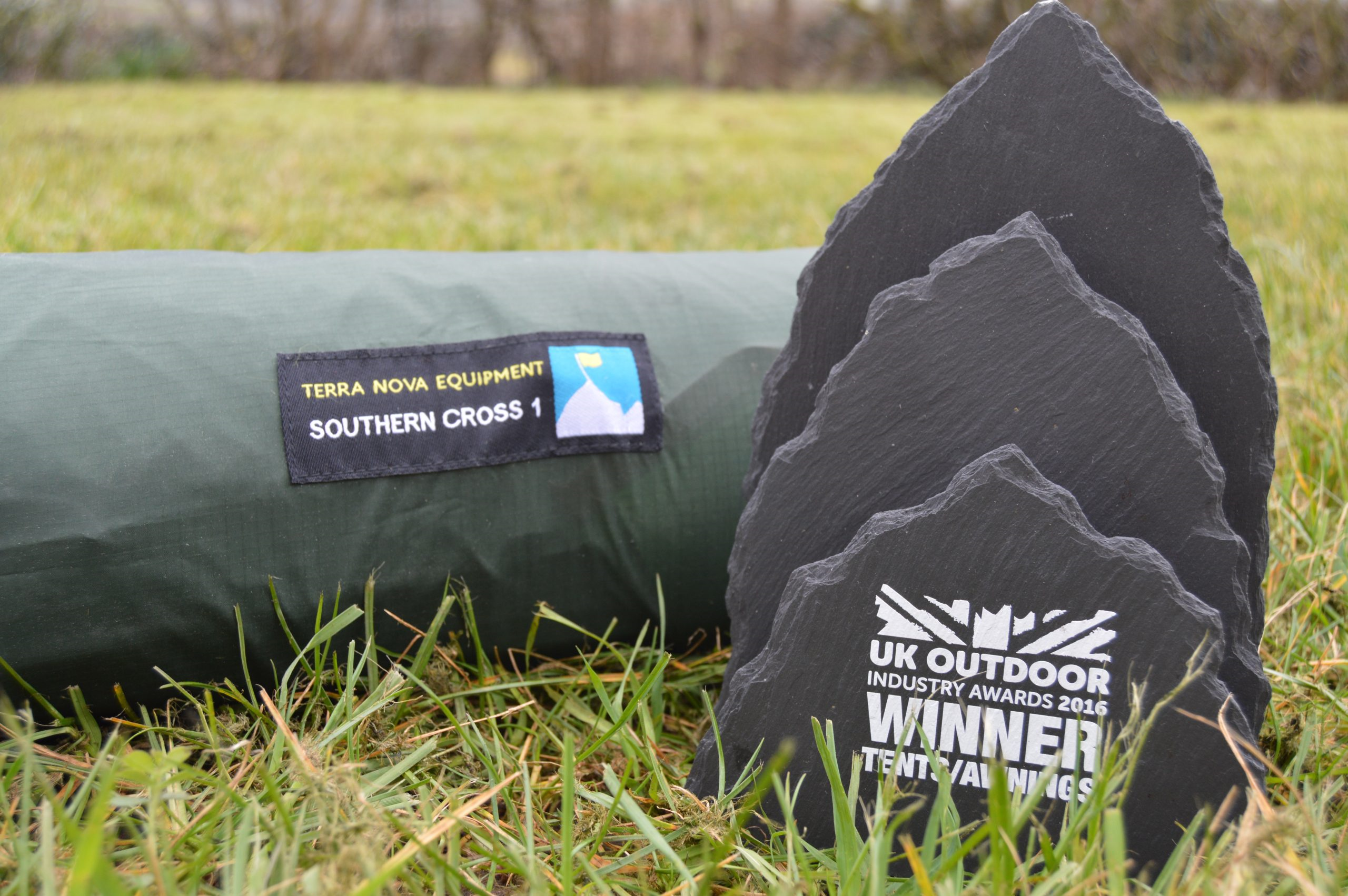 Green rolled Terra Nova Equipment 'Southern Cross 1' tent bag resting on grass beside three stacked black slate awards, the front slate displaying a white 'UK Outdoor Industry Awards 2016 WINNER' logo.
