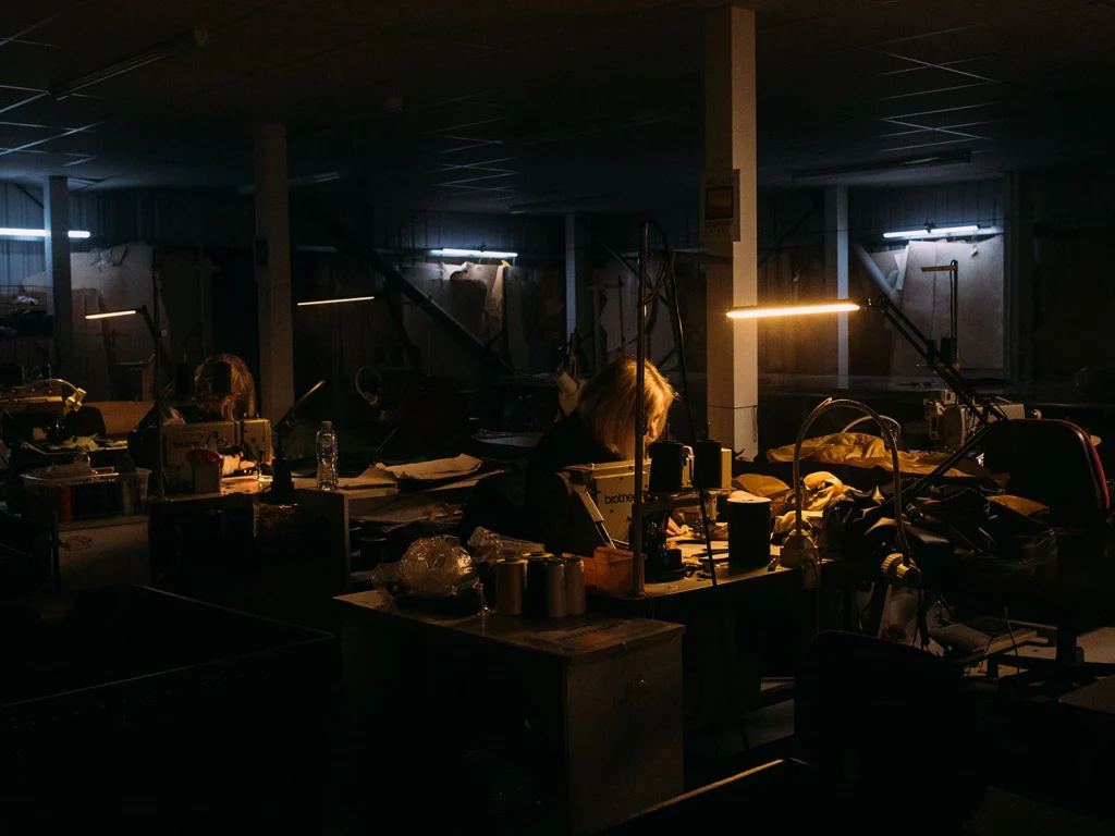 Dimly lit garment workshop with a person hunched over a Brother sewing machine under a warm task lamp, surrounded by piles of fabric, thread spools and industrial equipment with cool ceiling fluorescents in the background.