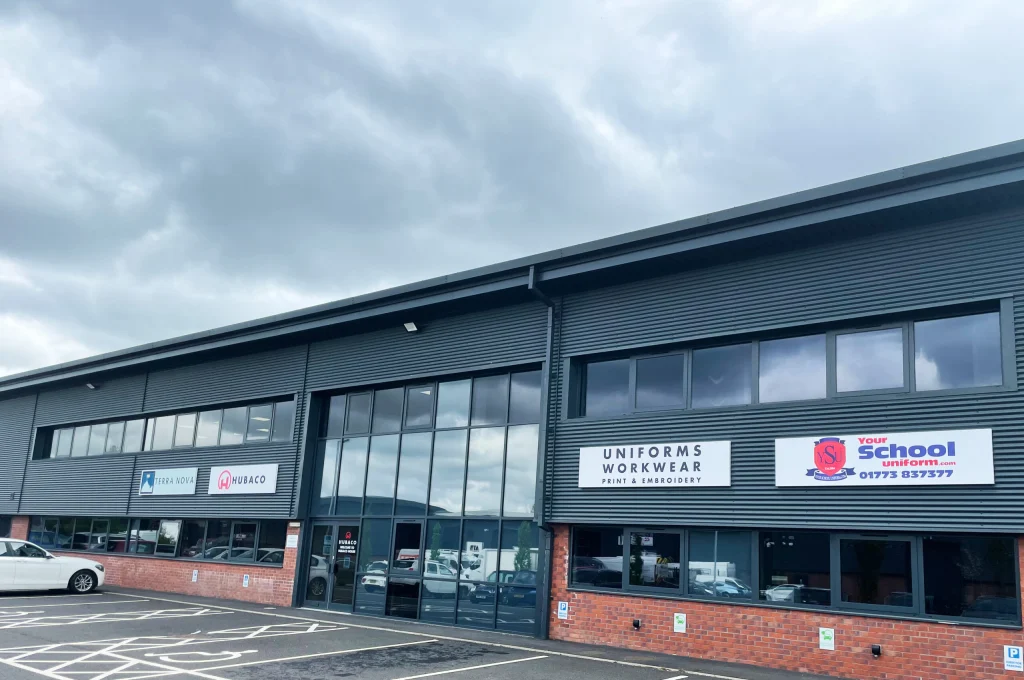 Exterior of a modern grey industrial building with a large glass entrance, brick lower wall and parking bays in front, under a cloudy sky, showing signs for Uniforms Workwear Print & Embroidery, Your School Uniform and other business logos.