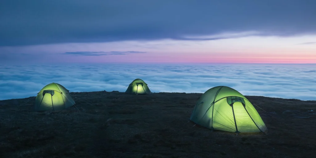 Three glowing green tents—one in the foreground right, one left, and one centered further back—pitched on a grassy ridge above a vast sea of clouds at dusk beneath a pastel pink and blue sky.