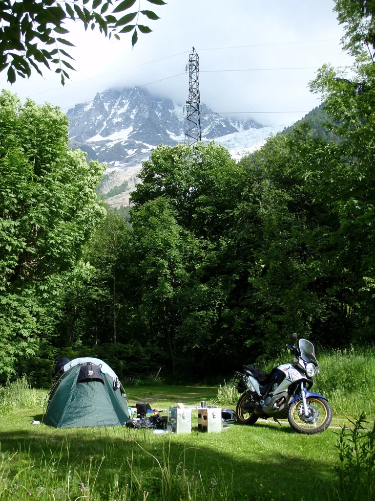 A green dome tent and scattered camping gear beside a parked adventure motorcycle on a sunlit grassy clearing, surrounded by dense trees with a partly cloud‑shrouded snow‑capped mountain and a transmission tower in the background.