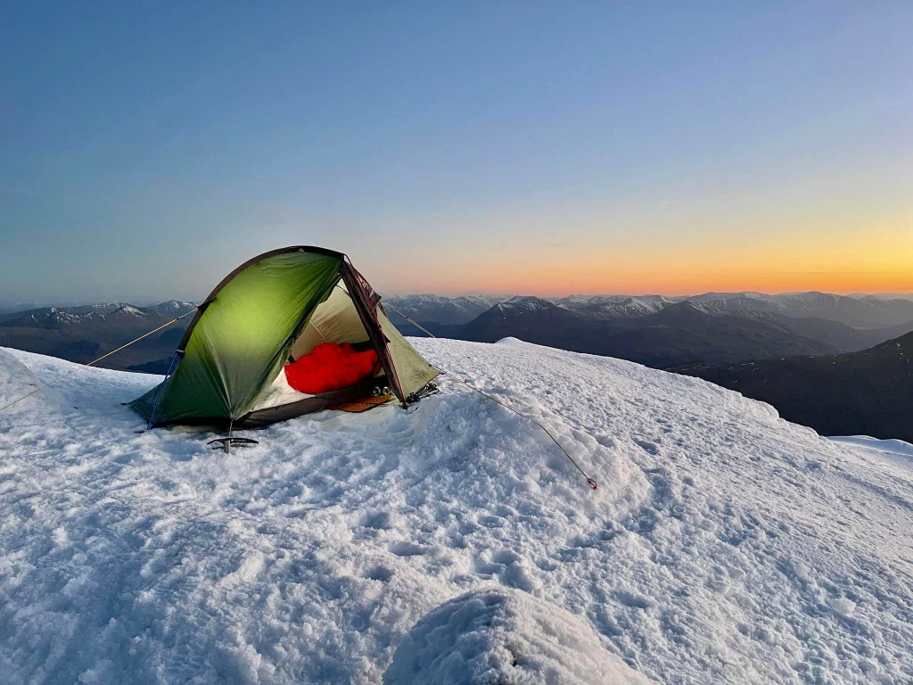 ls_Mark-Dack_snow_mountian_2022 Green two-person tent pitched on a snowy mountain summit at sunrise, a red sleeping bag visible inside, guy lines and an ice axe anchoring it, with distant snow-capped peaks under a clear orange-blue sky.